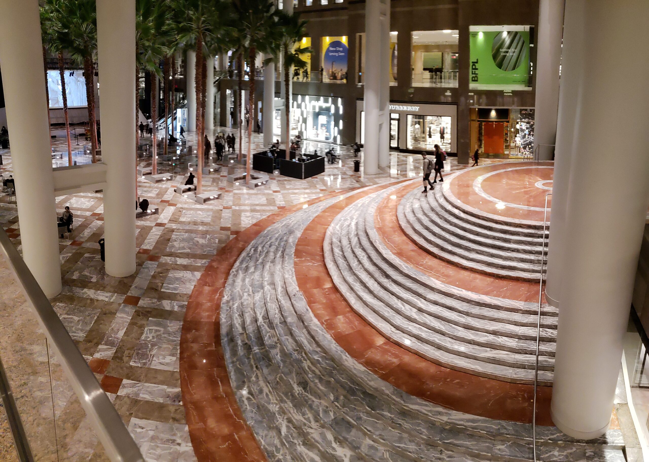 A fancy plaza inside a large building in NYC with a large, half-circle staircase and palm trees with a skylit roof.